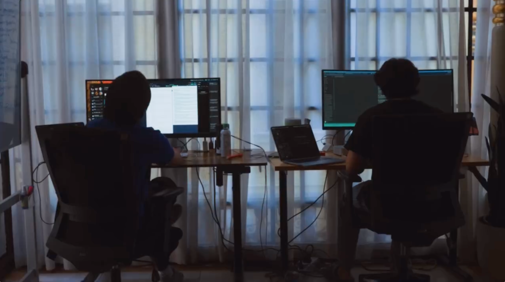 Two people working at desktop computers beside tall windows.
