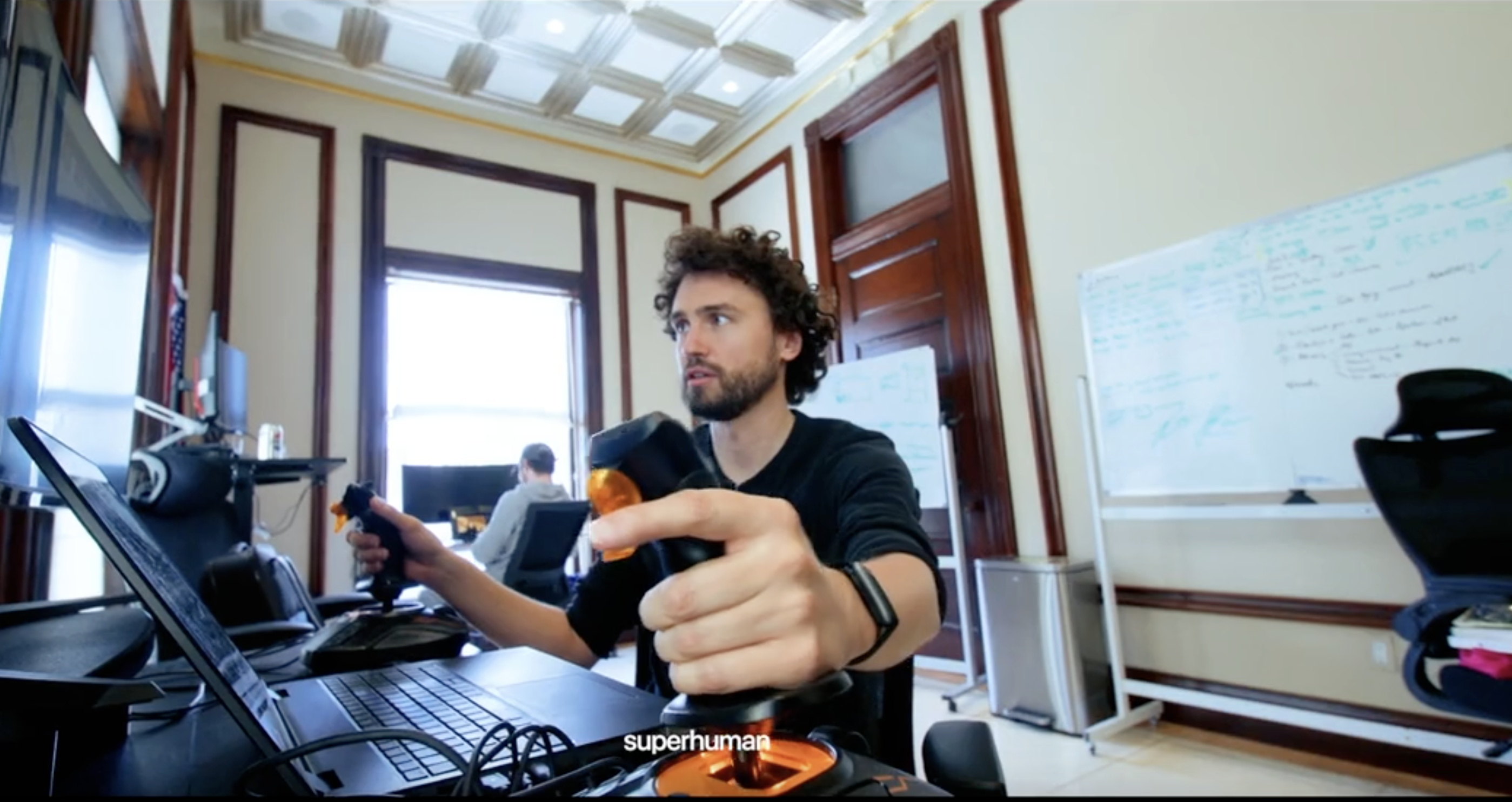 A founder with curly hair using flight-simulator joysticks at a laptop in an ornate mansion office, with whiteboards behind him.